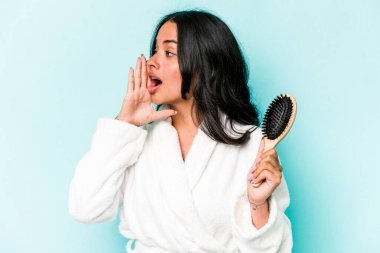 Mavi arka planda izole edilmiş saç fırçası tutan genç İspanyol kadın bağırıyor ve avuç içini açık tutuyor..