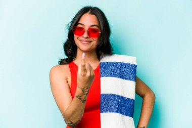 Mavi arka planda izole edilmiş plaj havlusu tutan genç İspanyol kadın sanki seni davet ediyormuş gibi seni işaret ediyor..