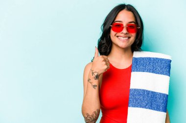 Mavi arka planda izole edilmiş plaj havlusu tutan genç İspanyol kadın gülümsüyor ve başparmağını kaldırıyor.