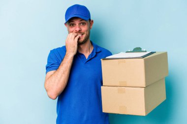 Kafkasyalı genç teslimatçı mavi arka planda izole edilmiş tırnak yiyor, gergin ve çok endişeli..