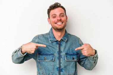 Beyaz arka planda yaşayan beyaz tenli genç bir adam gömlek fotokopi alanını gösteriyor, gururlu ve kendinden emin.