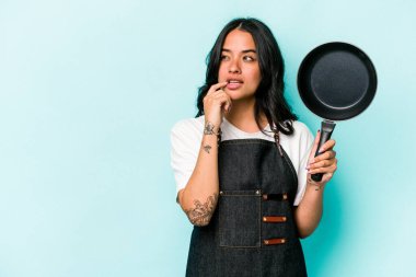Genç İspanyol tencereli kadın elinde tavayla mavi arka planda izole edilmiş bir şekilde bir şeyin fotokopi odasına baktığını düşünerek rahatlıyor..