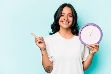 Mavi arka planda izole edilmiş bir saat tutan genç İspanyol kadın gülümsüyor ve boş alanda bir şey gösteriyor..