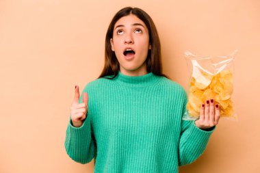 Latin kökenli genç bir kadın, elinde bej arka planda izole edilmiş bir paket cipsle ağzı açık bir şekilde üst tarafı işaret ediyor..