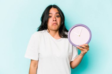 Mavi arka planda izole edilmiş bir saat tutan genç İspanyol kadın omuzlarını silker ve gözleri açık kalır..