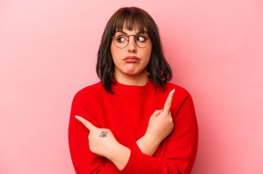 Pembe arka plan noktalarında izole edilmiş genç beyaz kadın iki seçenek arasında seçim yapmaya çalışıyor..