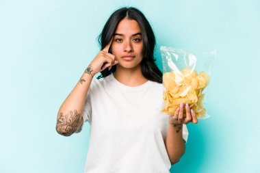Mavi arka planda bir çanta dolusu cipsi elinde tutan genç İspanyol kadın şakağı parmağıyla işaret ediyor, düşünüyor ve bir göreve odaklanıyor..