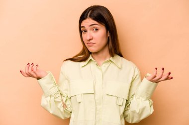 Latin kökenli genç bir kadın, bej arka planda izole edilmiş. Sorgulama sırasında omuz silkiyor ve şüphe duyuyor..