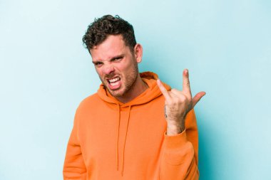 Mavi arka planda izole edilmiş genç beyaz adam parmaklarıyla kaya hareketi gösteriyor.