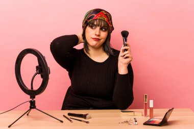 Makyaj yapan genç bir kadın pembe arka planda izole edilmiş bir makyaj dersi veriyor. Başının arkasına dokunuyor, düşünüyor ve bir seçim yapıyor..