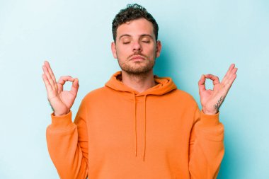 Mavi arka planda izole edilmiş genç beyaz adam yorucu bir günün ardından rahatlıyor. Yoga yapıyor..