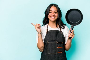 Genç İspanyol tencereli kadın elinde kızartma tavası tutarken mavi arka planda izole edilmiş gülümseyen ve işaret eden boş alanda bir şey gösteriyor..