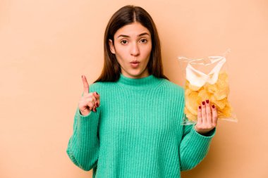 Latin kökenli genç bir kadın, elinde bej arka planda izole edilmiş bir çanta dolusu çip tutuyor. Harika bir fikri var, yaratıcılık kavramı..