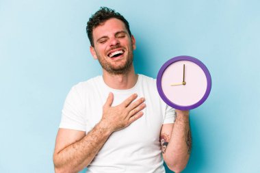 Mavi arka planda izole edilmiş bir saat tutan genç beyaz adam yüksek sesle gülüyor ve elini göğsünde tutuyor..