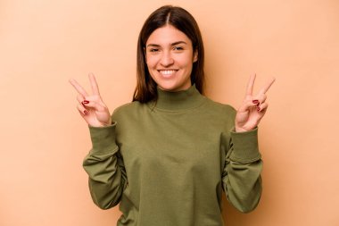 Bej arka planda izole edilmiş genç İspanyol kadın zafer işareti gösteriyor ve gülümsüyor..
