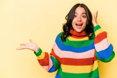 Sarı arka planda izole edilmiş genç İspanyol kadın avuç içinde fotokopi alanı tutuyor, el üstünde tutuyor. Şaşırmış ve mutlu..