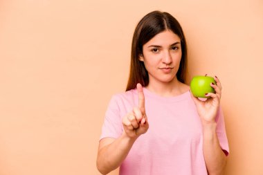 Elinde bej arka planda izole edilmiş bir elma tutan genç İspanyol kadın bir numarayı parmağıyla gösteriyor..
