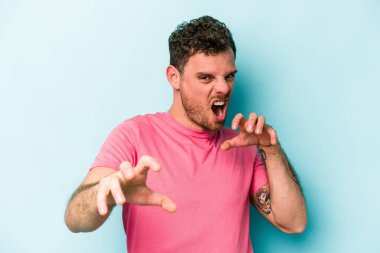 Mavi arka planda izole edilmiş genç beyaz adam pençelerini kedi taklidi yaparken gösteriyor, agresif bir hareket..