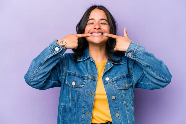 Mor arka planda izole edilmiş genç İspanyol kadın gülümsüyor, parmaklarını ağıza doğrultuyor..