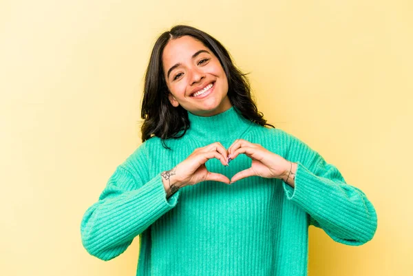 Sarı arka planda izole edilmiş genç İspanyol kadın gülümsüyor ve elleriyle kalp şeklini gösteriyor..