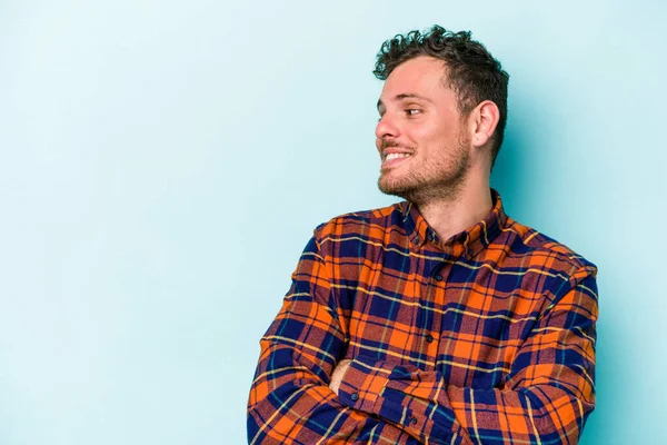 Mavi arka planda izole edilmiş genç beyaz adam kollarını kavuşturarak gülümsüyor..