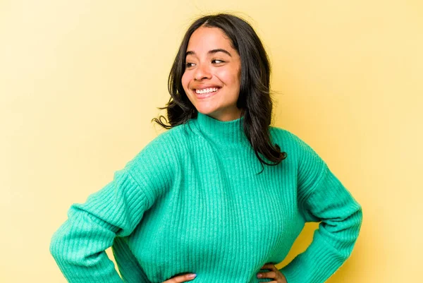 Sarı arka planda izole edilmiş genç İspanyol kadın mutlu bir şekilde gülüyor ve ellerini karnının üstünde tutarak eğleniyor..