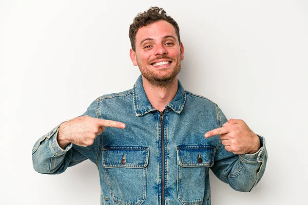 Beyaz arka planda yaşayan beyaz tenli genç bir adam gömlek fotokopi alanını gösteriyor, gururlu ve kendinden emin.