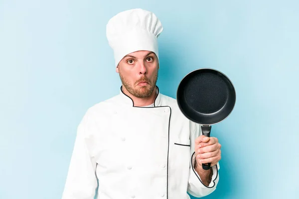 年轻的高加索厨师 拿着被蓝色背景隔离的飞盘 耸了耸肩 睁开眼睛迷惑不解 — 图库照片