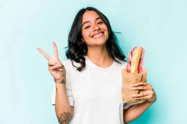 年轻的惊慌失措的女人吃着一个蓝色背景的三明治 快乐而无忧无虑 用手指展示着和平的象征 — 图库照片