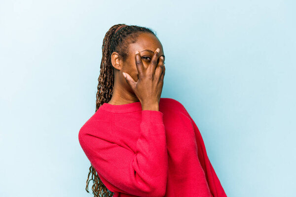 Young african american woman isolated on blue background blink at the camera through fingers, embarrassed covering face.