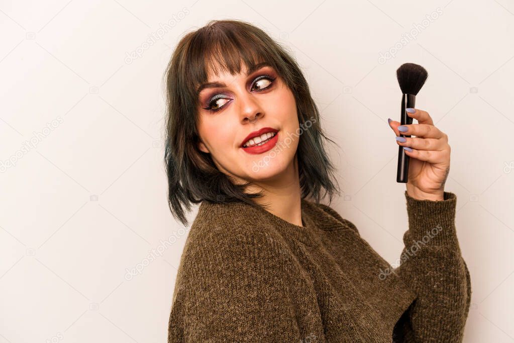 Young caucasian makeup artist woman holding a makeup brush isolated on white background looks aside smiling, cheerful and pleasant.