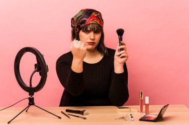 Makyaj yapan genç bir kadın pembe arka planda izole edilmiş makyaj dersi veriyor. Kameraya yumruk, agresif yüz ifadesi gösteriyor..