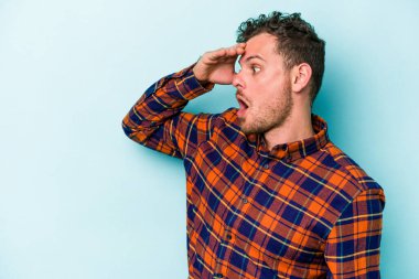 Kafkasyalı genç adam mavi arka planda izole edilmiş, uzaklara bakıyor ve elini alnına koyuyor..