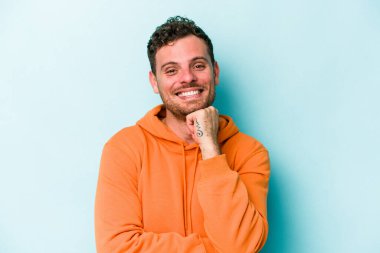Mavi arka planda izole edilmiş genç beyaz adam gülümsüyor ve kendinden emin, elleriyle çenesine dokunuyor..