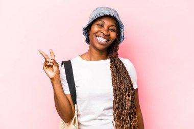 Genç Afrikalı Amerikalı bir kadın pembe arka planda izole edilmiş bir şekilde sahile gidiyor neşeli ve tasasız bir şekilde parmaklarıyla barış sembolü gösteriyor..