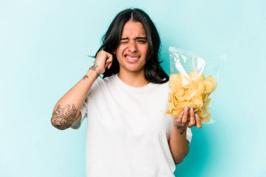 Mavi arka planda izole edilmiş bir paket cipsi tutan genç İspanyol kadın elleriyle kulaklarını kapatıyor..