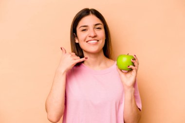 Elinde bej arka planda izole edilmiş bir elma tutan genç İspanyol kadın parmaklarıyla bir cep telefonu hareketi gösteriyor..