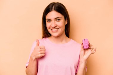 Latin kökenli genç bir kadın, elinde bej arka planda izole edilmiş bir araba tutuyor gülümsüyor ve başparmağını kaldırıyor.