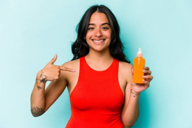 Mavi arka planda izole edilmiş güneş kremi tutan genç İspanyol kadın gömlek fotokopi alanını işaret ediyor, gururlu ve kendinden emin.