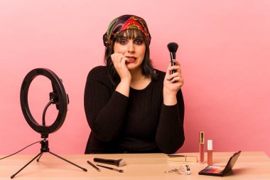 Makyaj eğitmeni genç bir kadın pembe arka planda izole edilmiş tırnak yiyor, gergin ve endişeli..