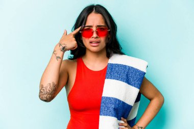 Mavi arka planda izole edilmiş plaj havlusu tutan genç İspanyol kadın işaret parmağıyla yaptığı hayal kırıklığı jestini gösteriyor..