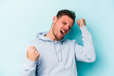 Kafkasyalı genç adam mavi arka planda izole edilmiş kaygısız ve heyecanlı tezahüratlar yapıyor. Zafer konsepti.