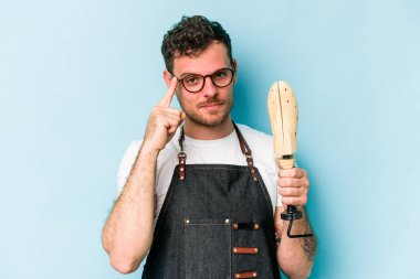 Kafkasyalı genç kunduracı, mavi arka planda izole edilmiş. Şakağı işaret ediyor, düşünüyor, bir göreve odaklanıyor..