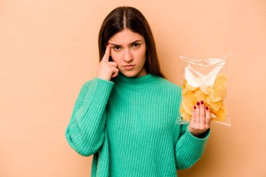 Latin kökenli genç bir kadın, elinde bej arka planda izole edilmiş bir çanta tutuyor. Şakağı işaret ediyor, düşünüyor, bir göreve odaklanıyor..