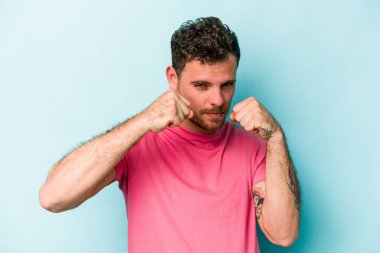 Kafkasyalı genç bir adam mavi arka planda izole edilmiş. Yumruk atıyor, sinirleniyor, kavga ediyor, kavga ediyor..