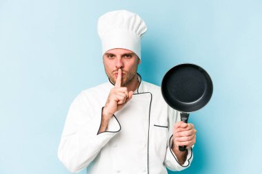 Kafkasyalı genç aşçı elinde uçan tavayla mavi arka planda izole edilmiş bir şekilde bir sır saklıyor ya da sessizlik istiyor..