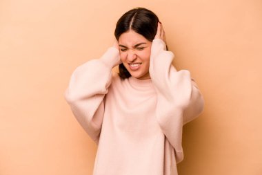 Latin kökenli genç kadın bej arka planda izole edilmiş kulakları elleriyle kapatıyor..