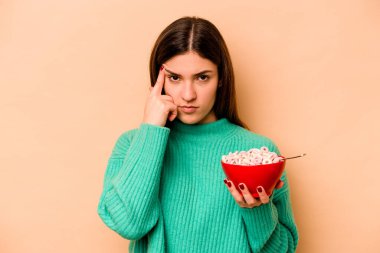 Latin kökenli genç bir kadın bej arka planda izole edilmiş mısır gevreği yiyor. Şakağı işaret ediyor, düşünüyor, bir göreve odaklanıyor..