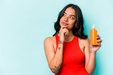 Mavi arka planda güneş kremi tutan genç İspanyol kadın şüpheli ve şüpheci bir ifadeyle yana bakıyor..