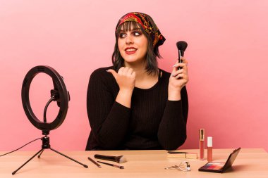 Makyaj eğitmeni genç bir kadın pembe arka plan noktalarına izole edilmiş, baş parmak uzakta, gülüyor ve kaygısız bir şekilde makyaj yapıyor..